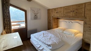 Une chambre à coucher avec un lit, une tête de lit en bois, une décoration murale représentant une tête de cerf, une fenêtre avec des rideaux et une vue sur un paysage montagneux.