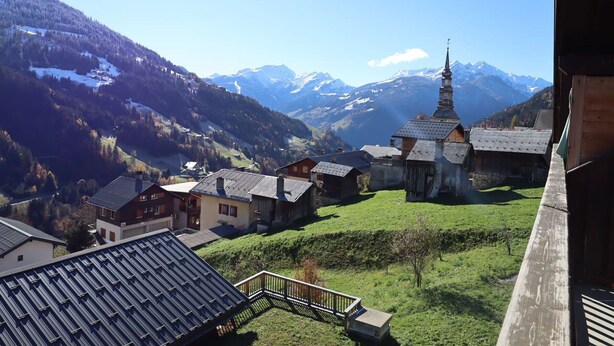 Un village aux maisons traditionnelles en bois, une église dotée d’un clocher imposant et, en arrière-plan, des montagnes enneigées.