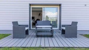 An outdoor patio with two grey wicker chairs and a bench, a sliding glass door leading to a room, and a view of the landscape.