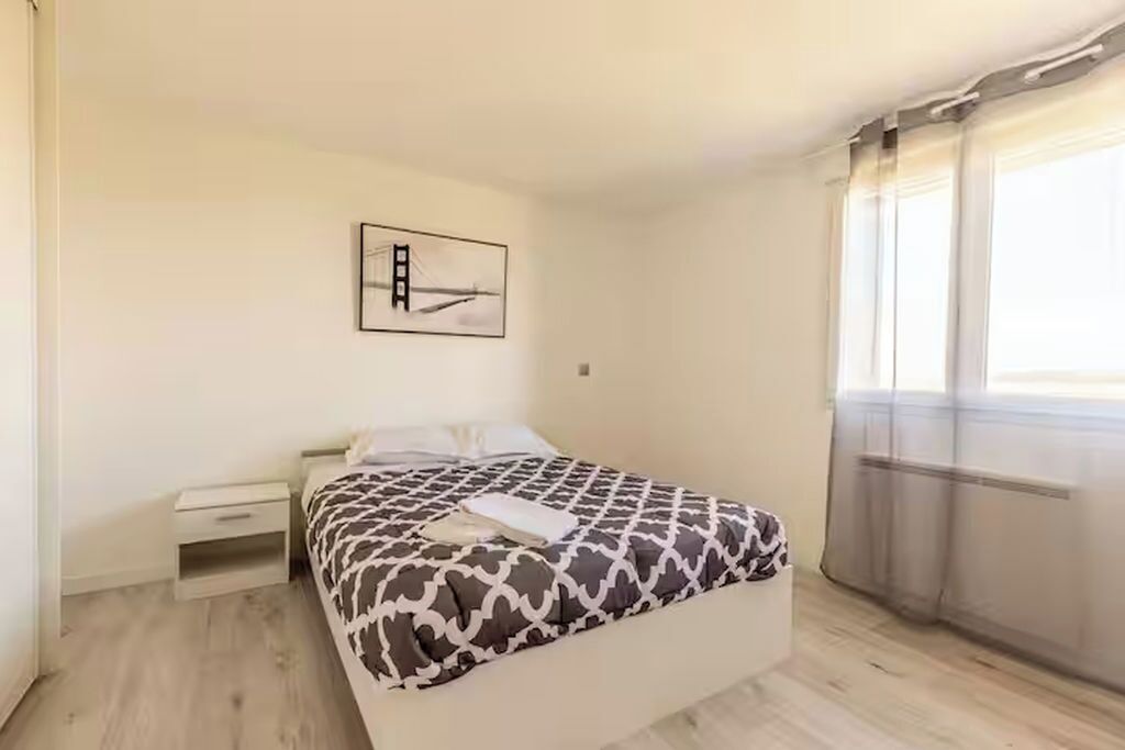 A bedroom with a bed, a nightstand, a small drawer, a framed picture on the wall, and a window with curtains.