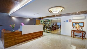 A hotel reception area with a wooden reception desk, a chandelier, a glass door leading to a restaurant, and a decorative table with a statue. - Hotel 91 Huda Premier (Gurugram)