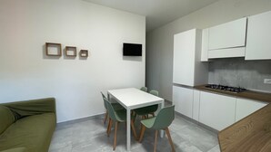 A compact kitchen with white cabinets, a gas stove, and a dining area with a white table and green chairs. - Renovated two-bedroom flat in Rosolina Mare (Rosolina Mare)