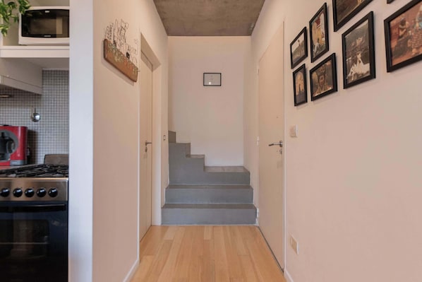 A narrow hallway with a staircase leading to a landing. There is a kitchen area on the left with a stove and a microwave. The walls are decorated with framed pictures.
