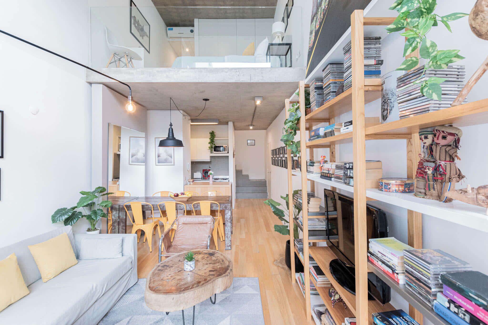 Amplia sala de estar con techo alto, comedor con sillas amarillas, sofá con cojines, mesa de centro y estantería llena de libros y objetos decorativos.