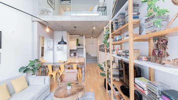 Amplia sala de estar con techo alto, comedor con sillas amarillas, sofá con cojines, mesa de centro y estantería llena de libros y objetos decorativos.