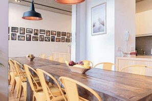 A dining area with a long wooden table, yellow chairs, and a fruit bowl. There are framed pictures on the wall and a kitchen area with a phone and a tiled backsplash.
