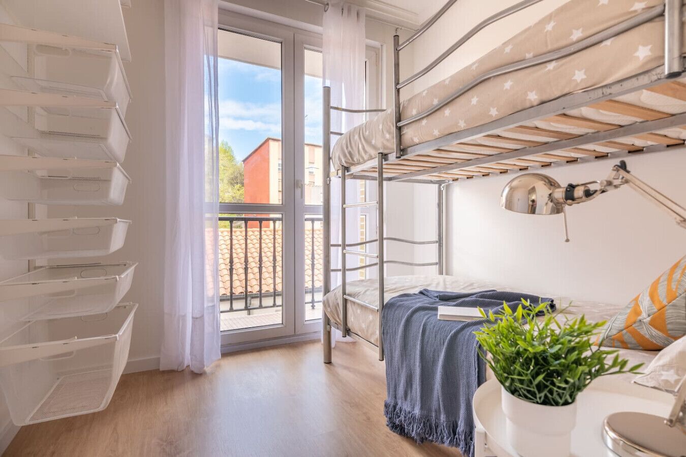 Een kamer in een studentencomplex met stapelbedden, een balkon, een bureau met lamp en een potplant.