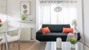 En moderne stue med en sort sofa, to orange puder, et glas sofabord, en hvid radiator, en hvid stol, et lille hvidt bord, en indrammet plante og lette gardiner.