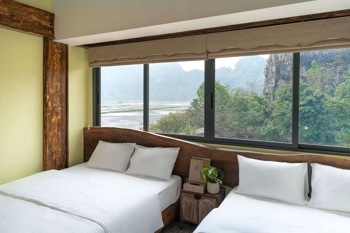 A hotel room with two beds, a large window with a view of a river and mountains, and a wooden headboard.