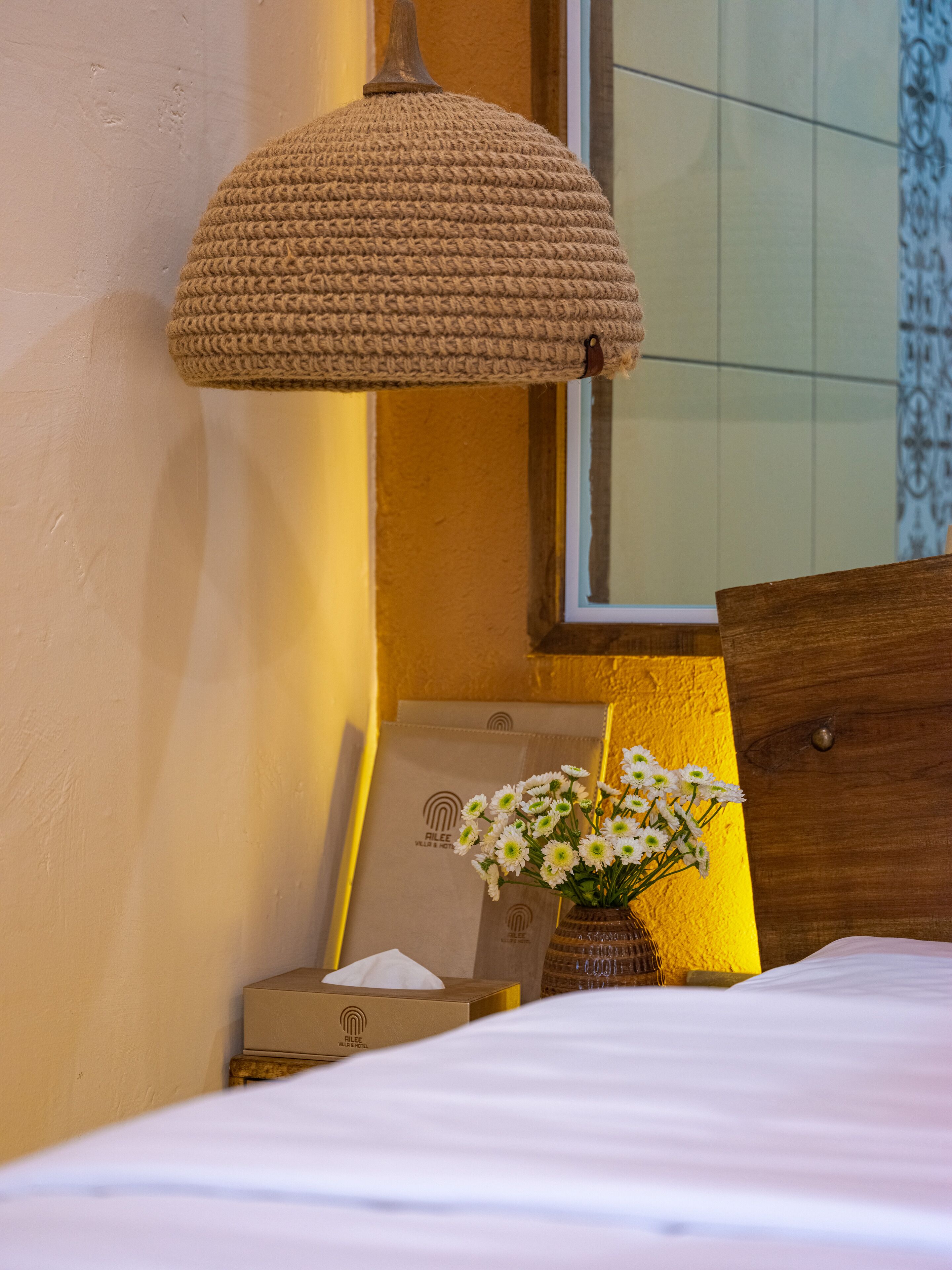 Een net opgemaakt bed met een witte deken, een geweven lamp, een vaas met witte bloemen en een ingelijst foto aan de muur.
