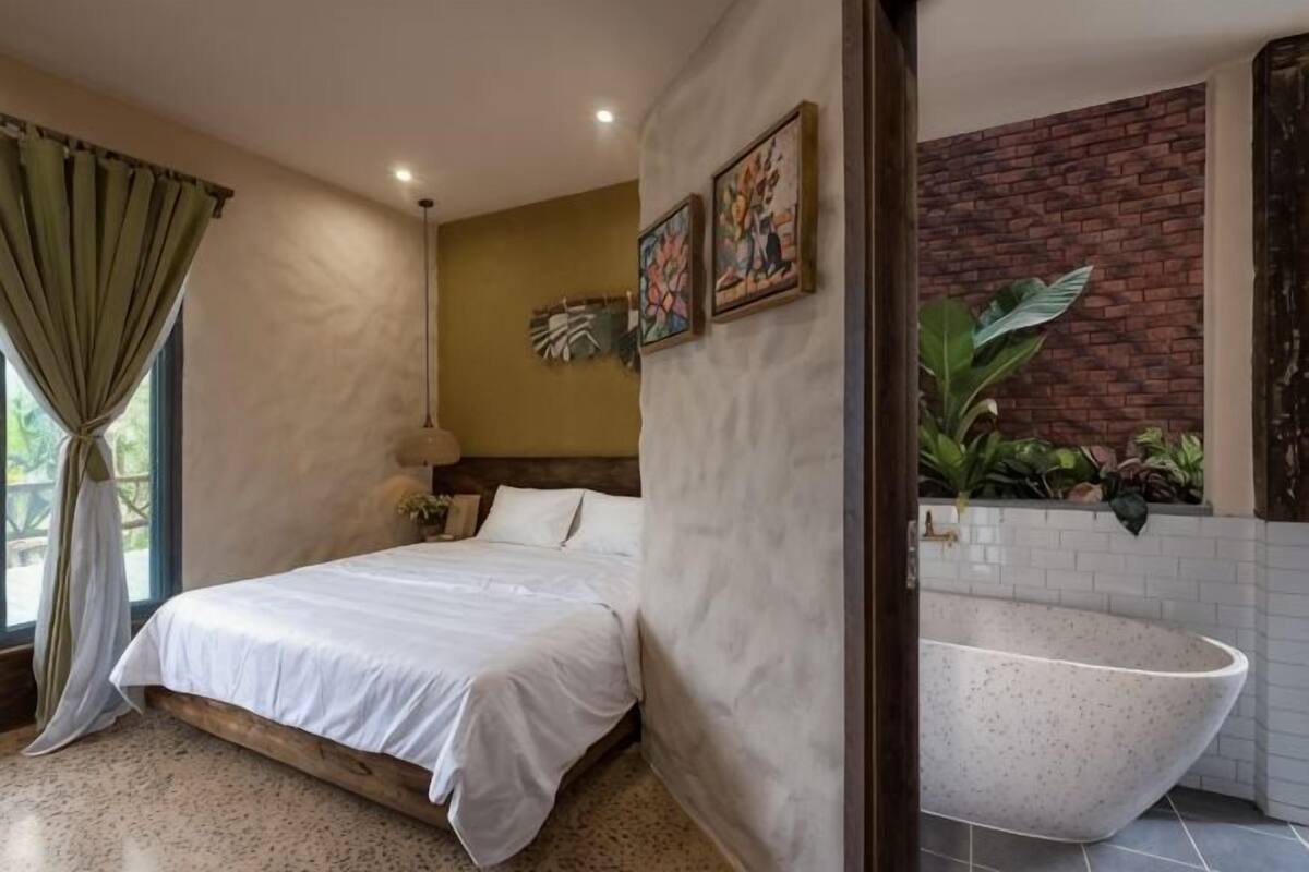 A bedroom with a bed, a large window with curtains, a wall-mounted light, and a bathroom with a bathtub.