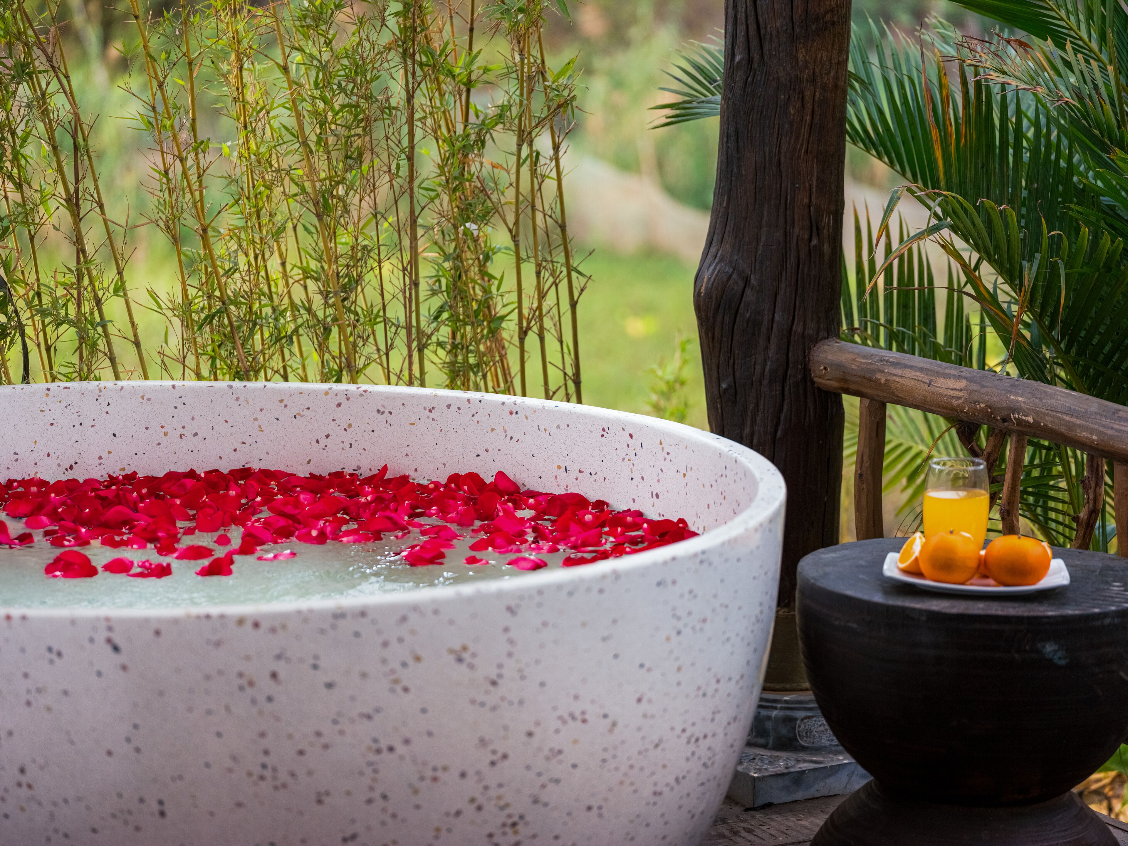 A spa setting with a large bowl filled with water and rose petals, a glass of orange juice, and a bowl of oranges on a wooden table.