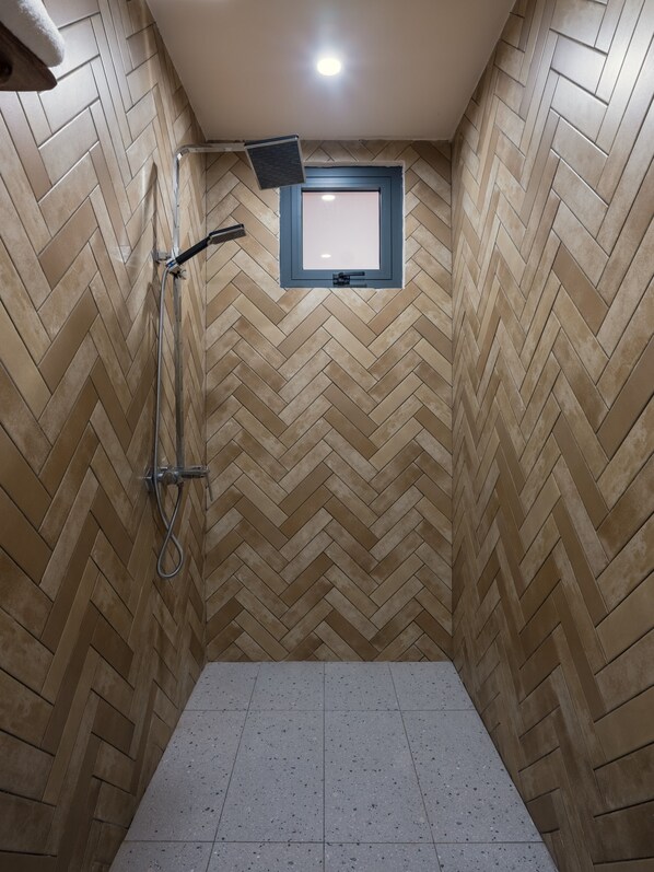 A shower room with a herringbone tile pattern on the walls and floor, a handheld showerhead, and a small window with a blue frame.