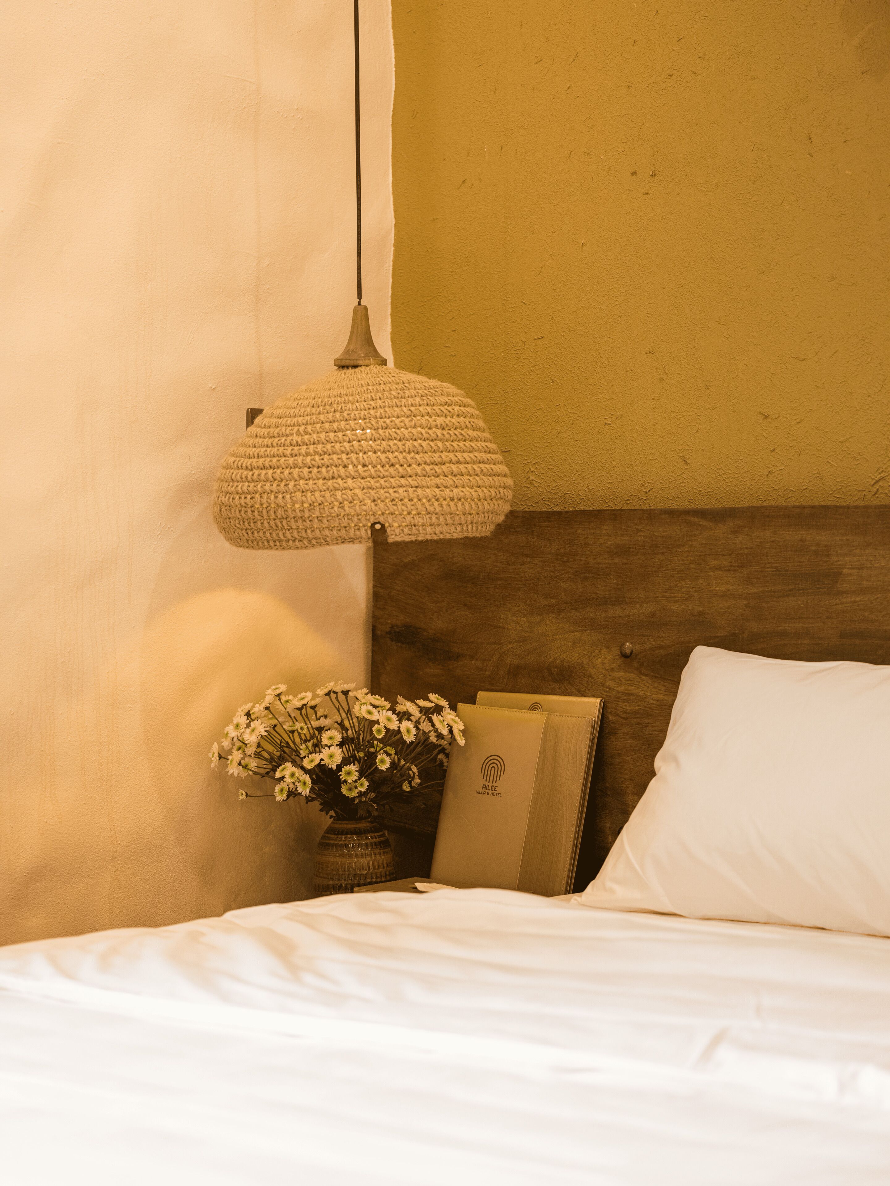 Un lit bien fait, avec du linge de lit blanc, une lampe de chevet, un vase rempli de fleurs et un livre posé sur la table de chevet.