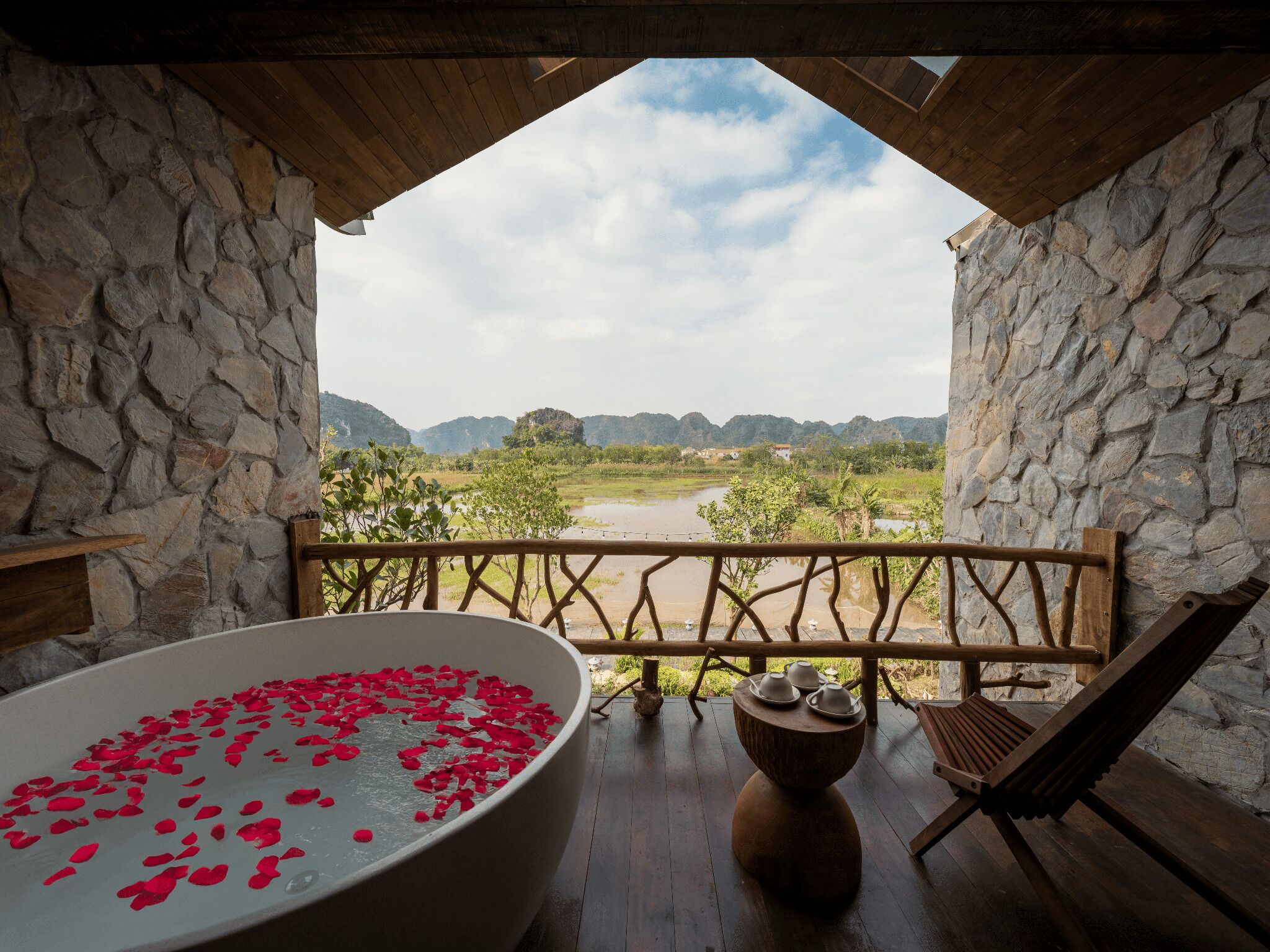 A stone-walled room with a large bathtub filled with rose petals, a wooden chair, and a view of a rural landscape.
