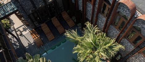 A rooftop pool area with wooden loungers, palm trees, and a stone wall.