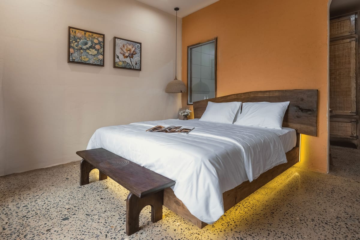 A bedroom with a large bed, a bench, two wall-mounted paintings, a mirror, and a textured floor.