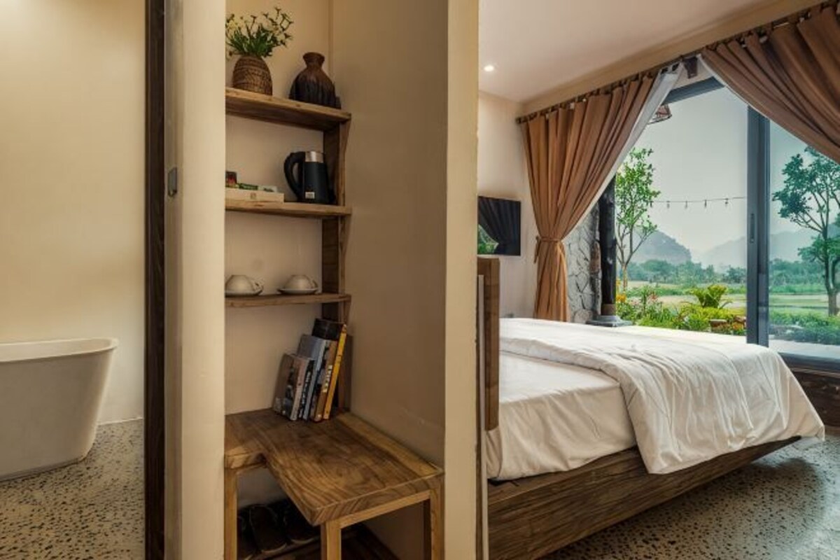 Une chambre avec un lit, une étagère en bois remplie de livres, une petite table, une fenêtre avec des rideaux et une vue sur la verdure.