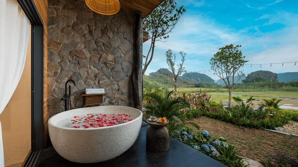 A spa area with a large freestanding bathtub filled with rose petals, a stone wall, and a view of a lush landscape. - AiLee Villa & Hotel Tamcoc (Nam Hoa Lu)