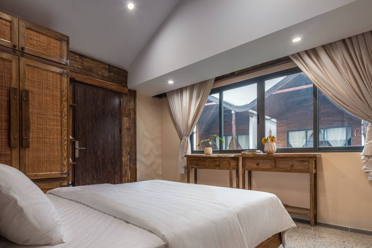 A bedroom with a large bed, wooden wardrobe, and a window with curtains.