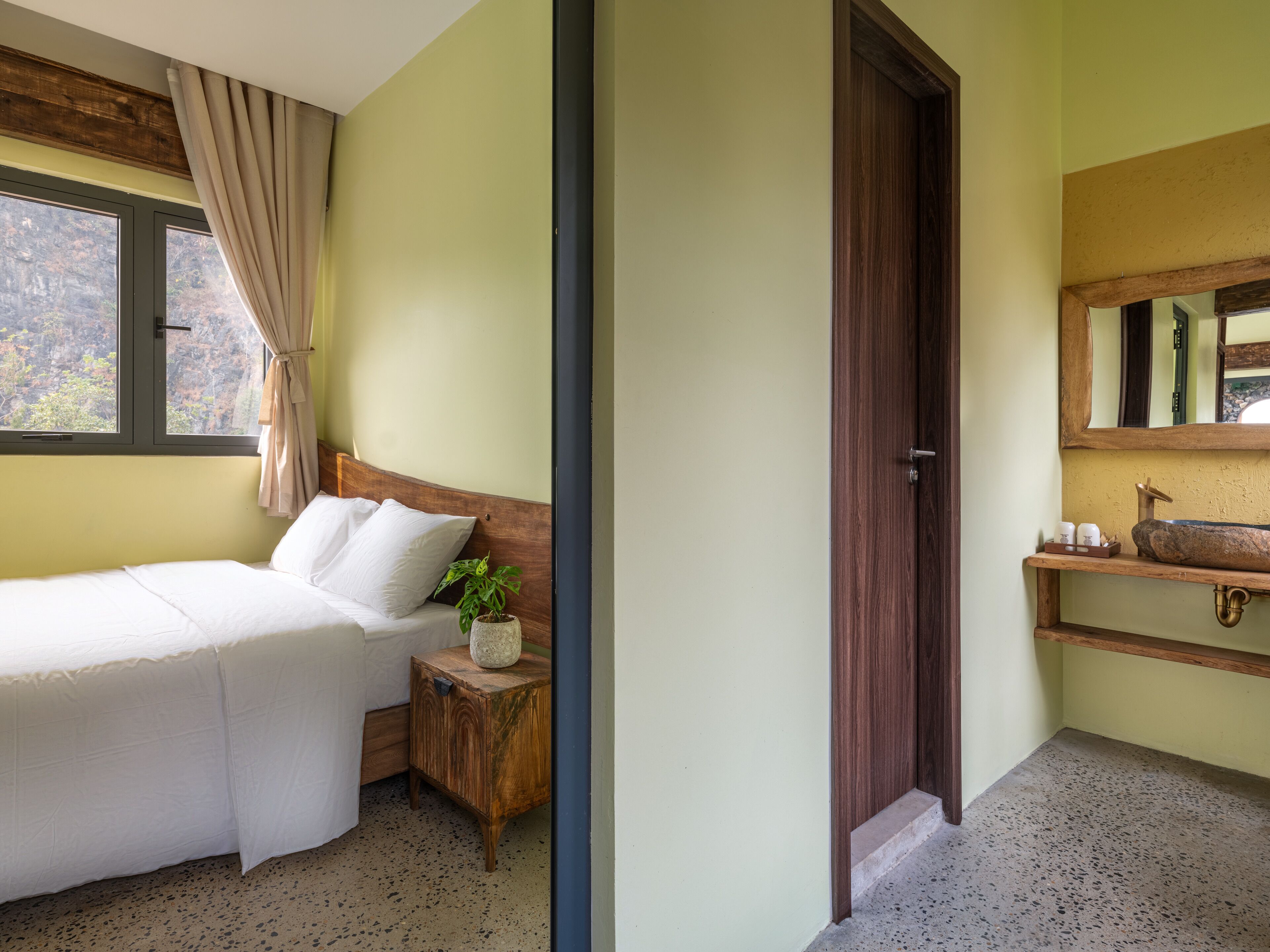 Een slaapkamer met een bed, een nachtkastje met een plant, een raam met gordijnen en een badkamer met een houten wastafel en spiegel.