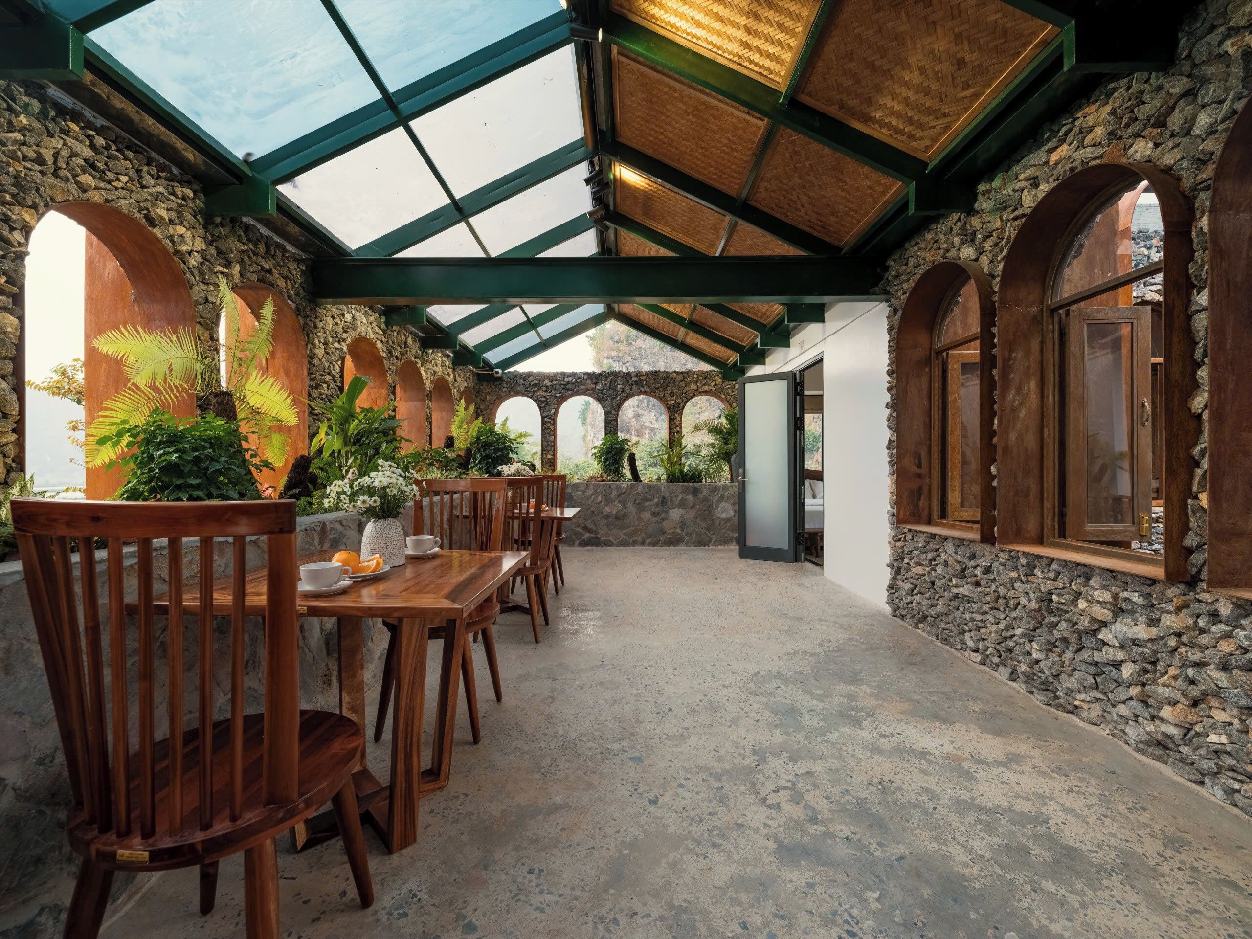 A covered patio with stone walls, wooden furniture, and a glass ceiling.