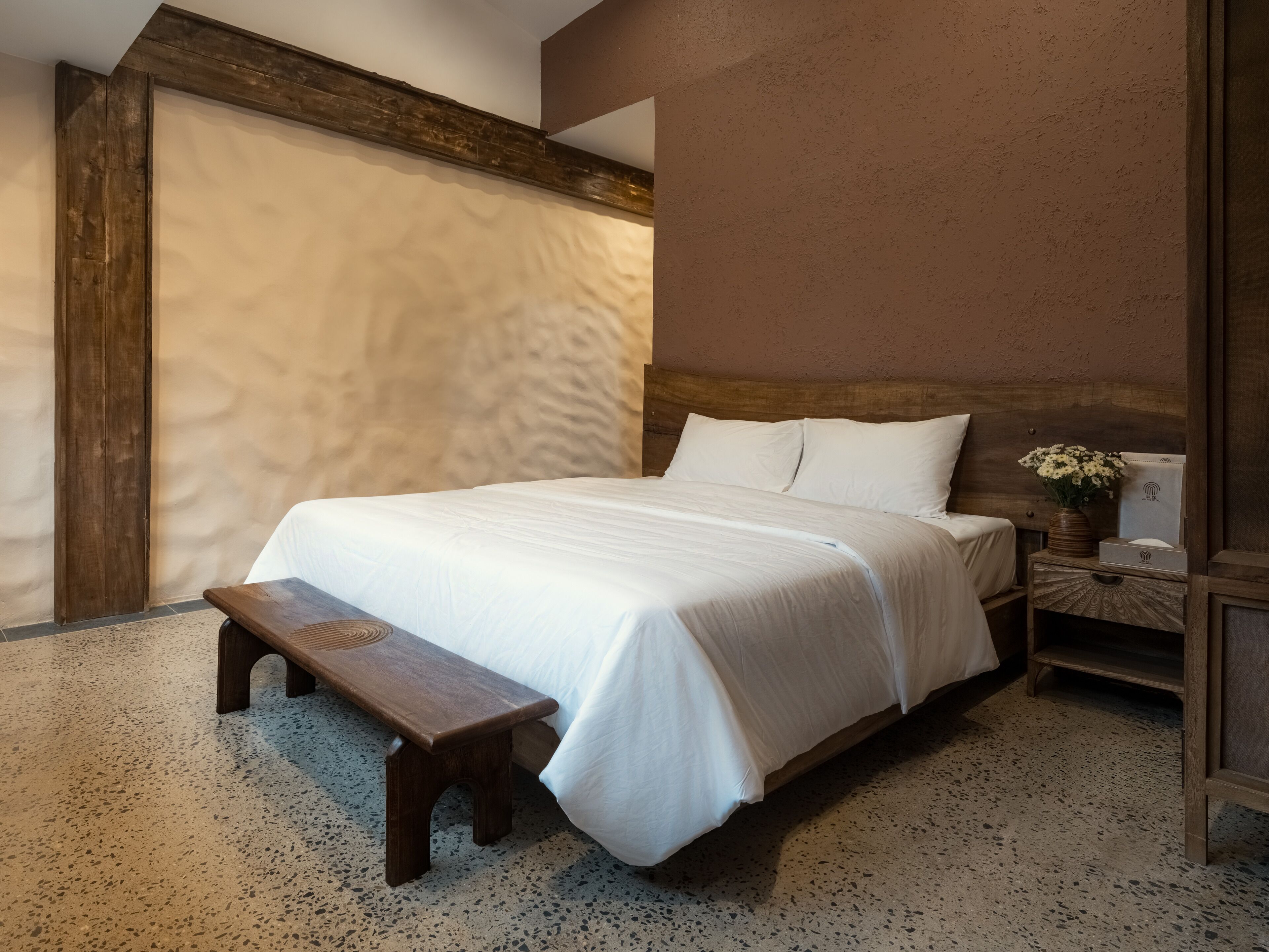 A bedroom with a large bed, a wooden bench, a nightstand with a vase of flowers, and a textured wall.