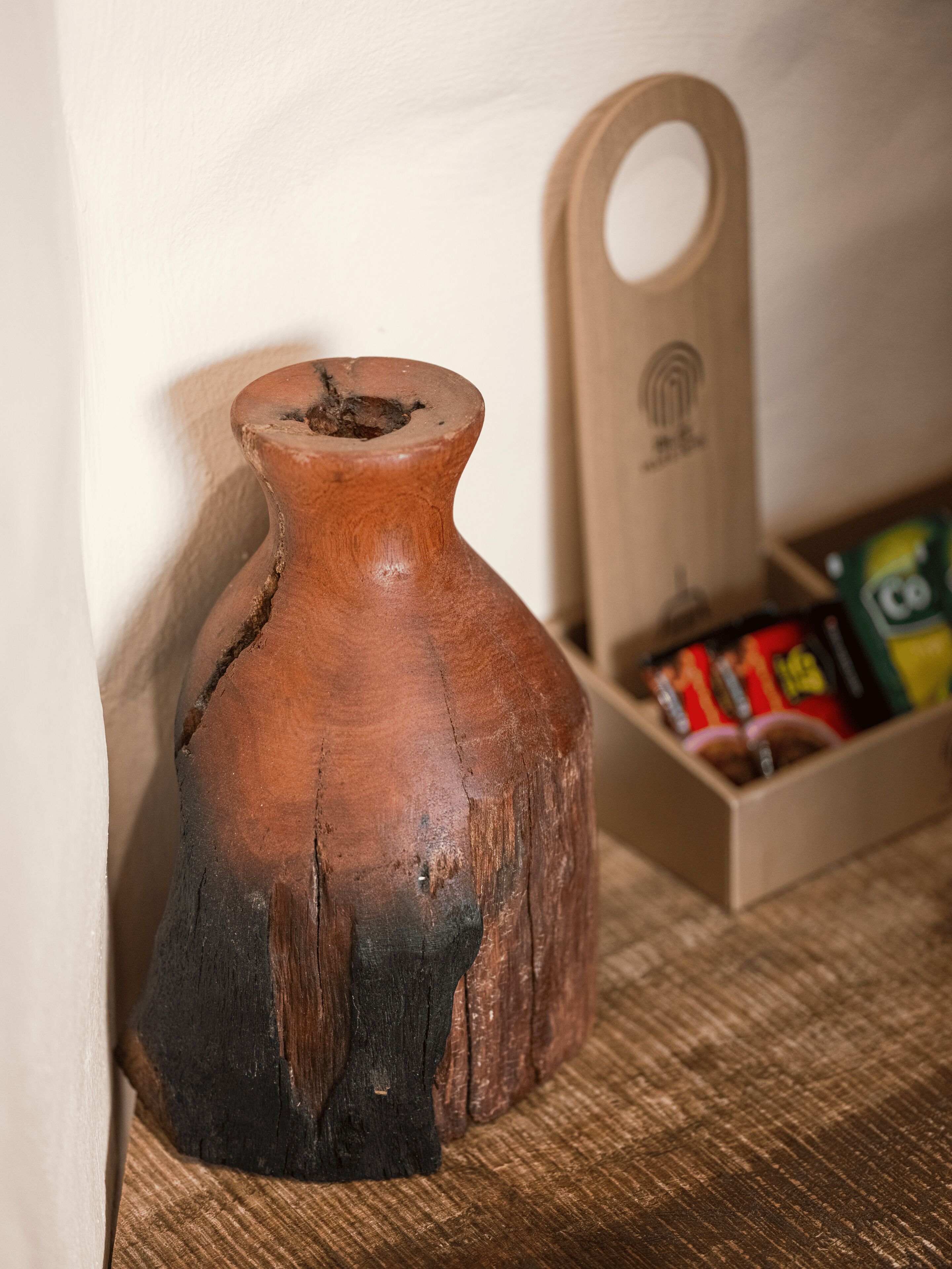 Un jarrón de madera rústico con asa, sobre una superficie tejida junto a una caja de madera con objetos de colores por dentro.