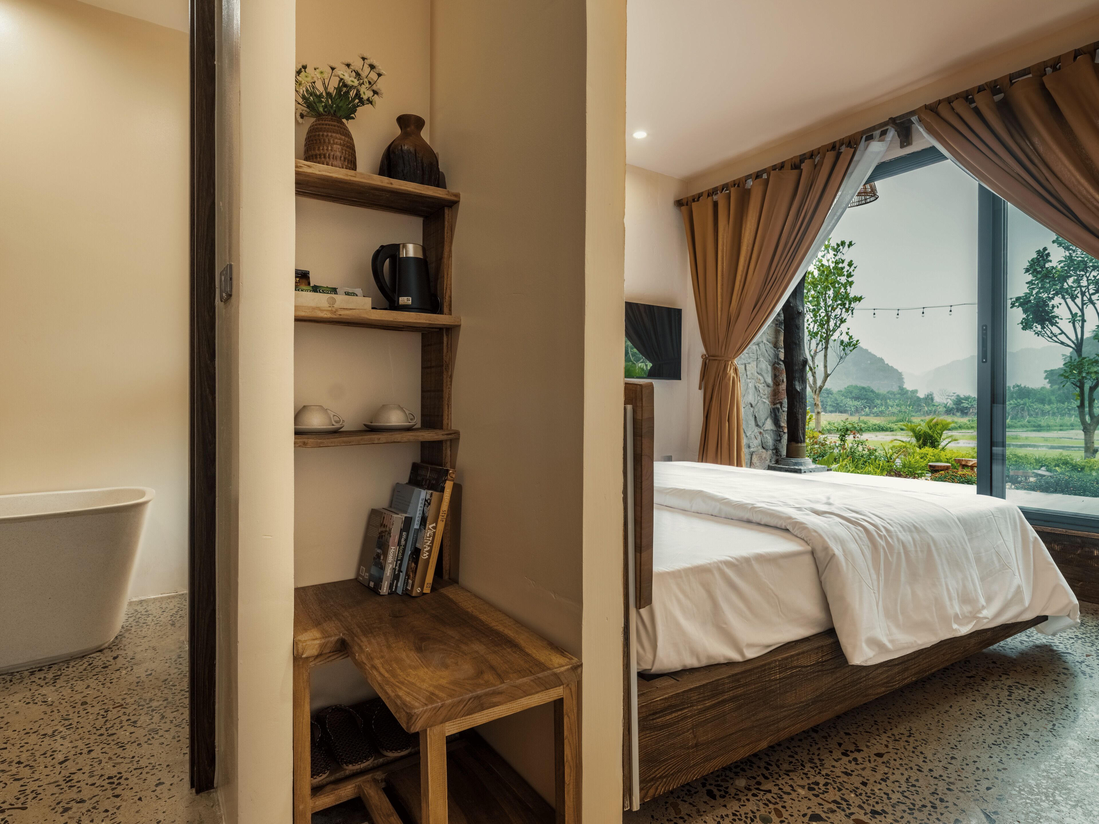 A bedroom with a bed, a wooden shelf with books, a coffee maker, and a view of greenery outside.