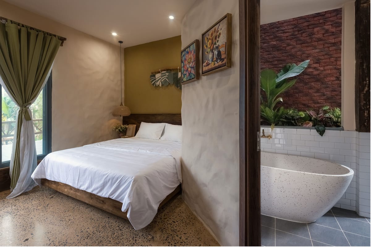 A bedroom with a large bed, a wall-mounted painting, a potted plant, and a view of a bathroom with a large bathtub.