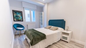 Um quarto com cama, uma cadeira, mesa de cabeceira, janela e radiador.