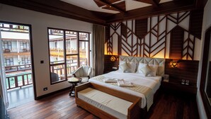 Ett hotellrum med en stor säng, en träbänk, en stol och utsikt över en traditionell byggnad genom stora fönster.