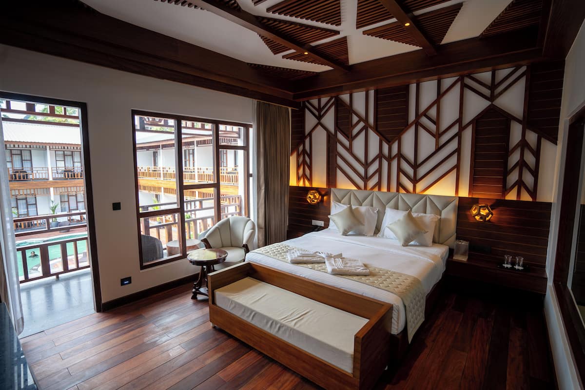 A hotel room with a large bed, a wooden bench, a chair, and a view of a traditional building through large windows.