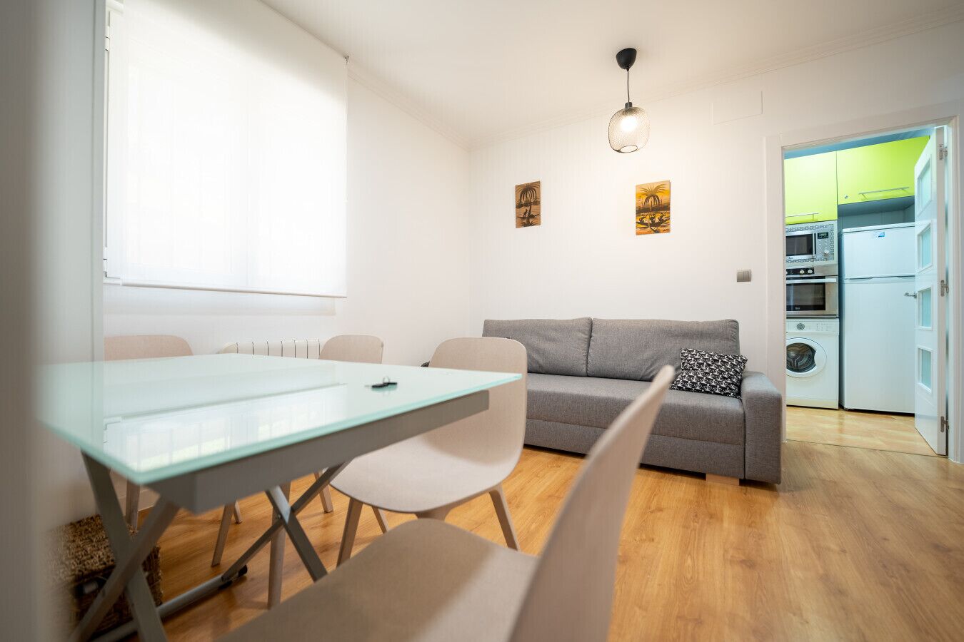 Een moderne woonkamer met een grijze bank, een glazen eettafel en een zichtbare keuken door een open deur.