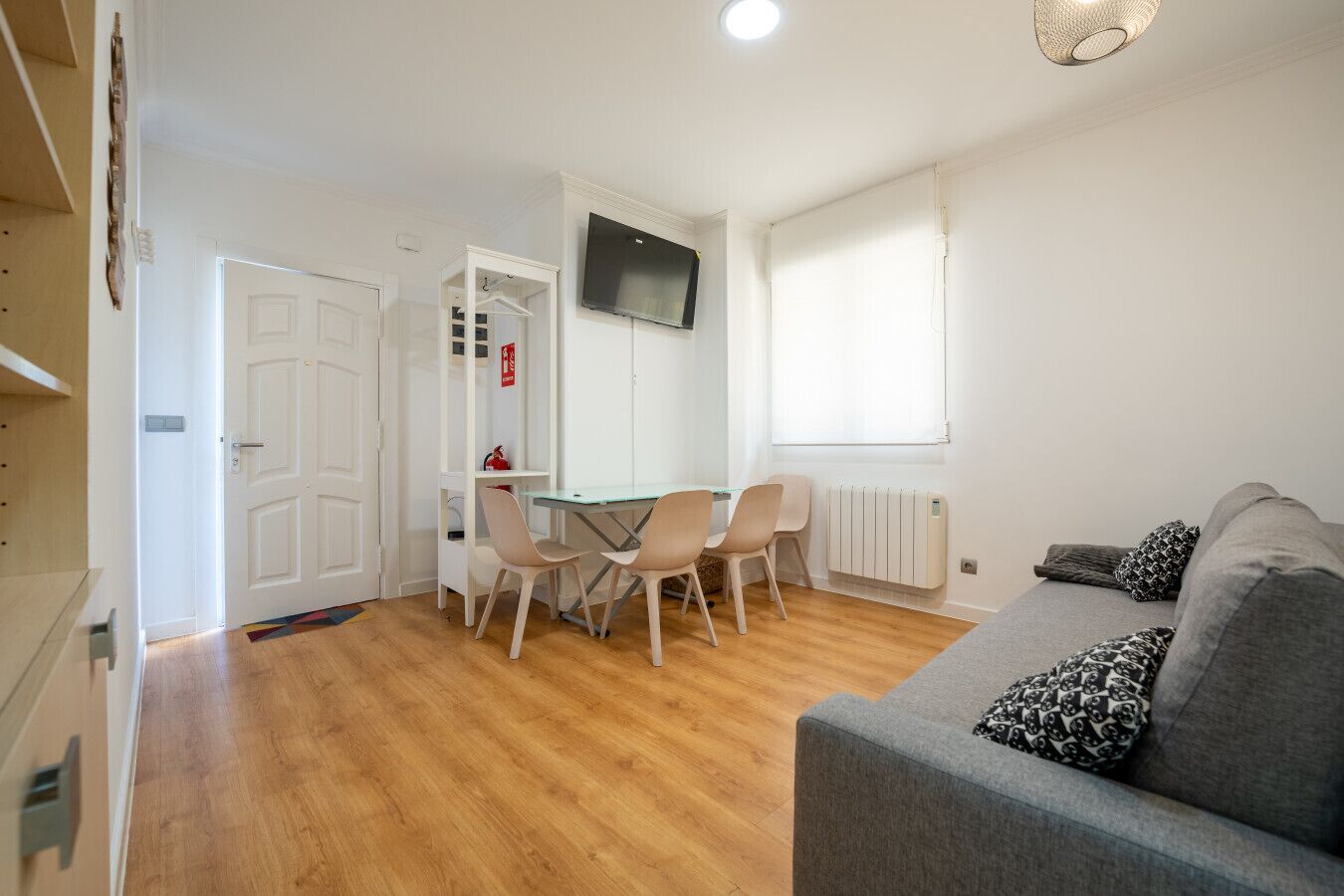 Een moderne woonkamer met een grijze bank, een eethoek met een tafel en stoelen, een aan de muur bevestigde televisie en een raam met zonwering.