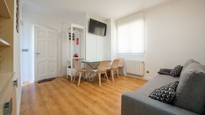 A modern living room with a grey sofa, a dining area with a table and chairs, a TV mounted on the wall, and a window with blinds. - Apto tranquilo y luminoso con cocina equipada (Santander)