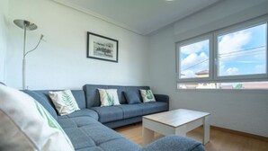 En moderne stue med en grå hjørnesofa, et hvidt sofabord og en gulvlampe. Et indrammet billede hænger på væggen, og der er et vindue med udsigt til himlen og bygninger.