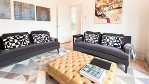Ein Wohnzimmer mit zwei Sofas, einem Couchtisch und einer Chandelieber. An den Wänden befinden sich abstrakte Gemälde und ein großes Poster.