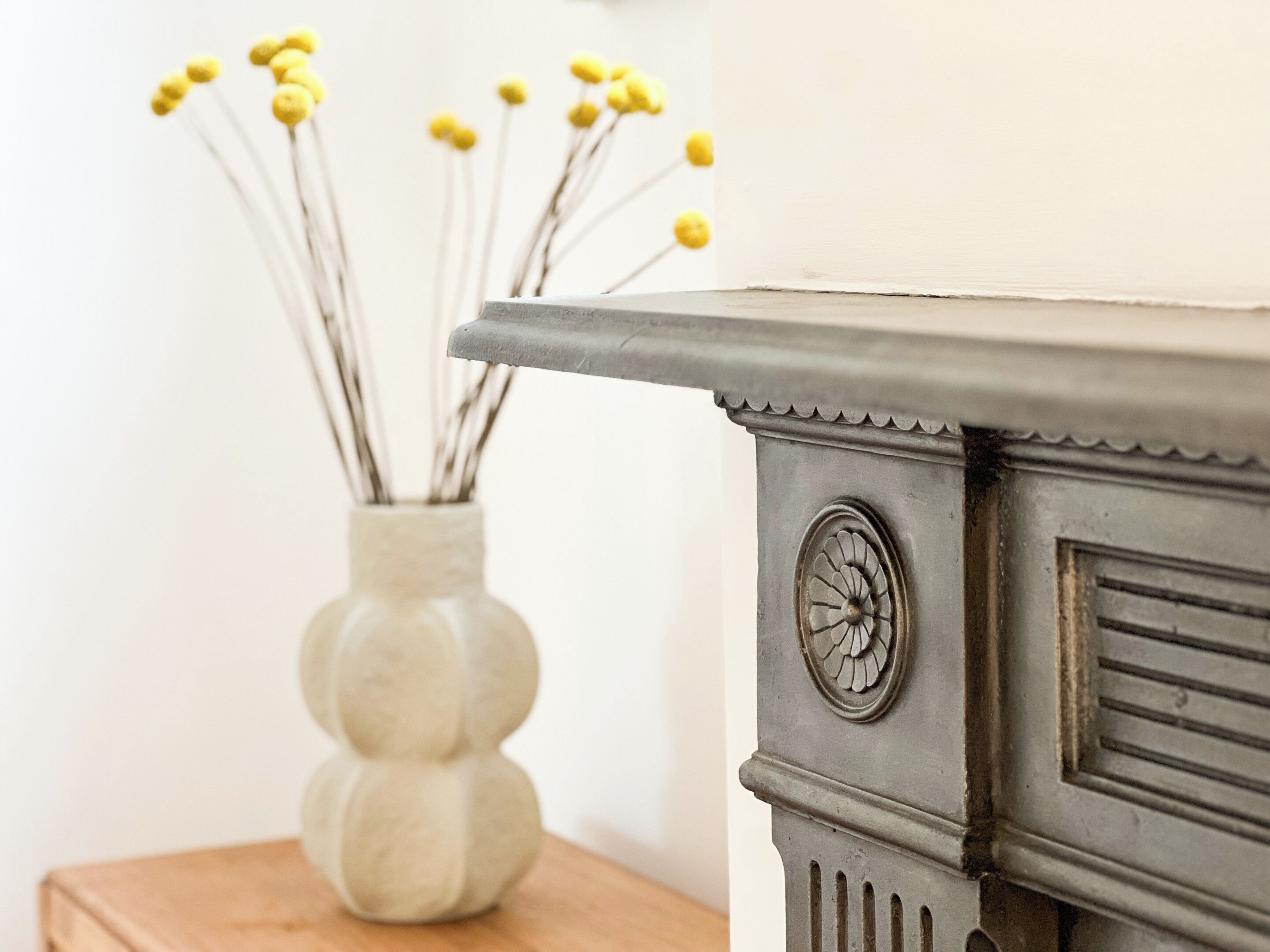 A decorative vase with yellow flowers on a wooden surface next to a fireplace with a decorative metal trim.
