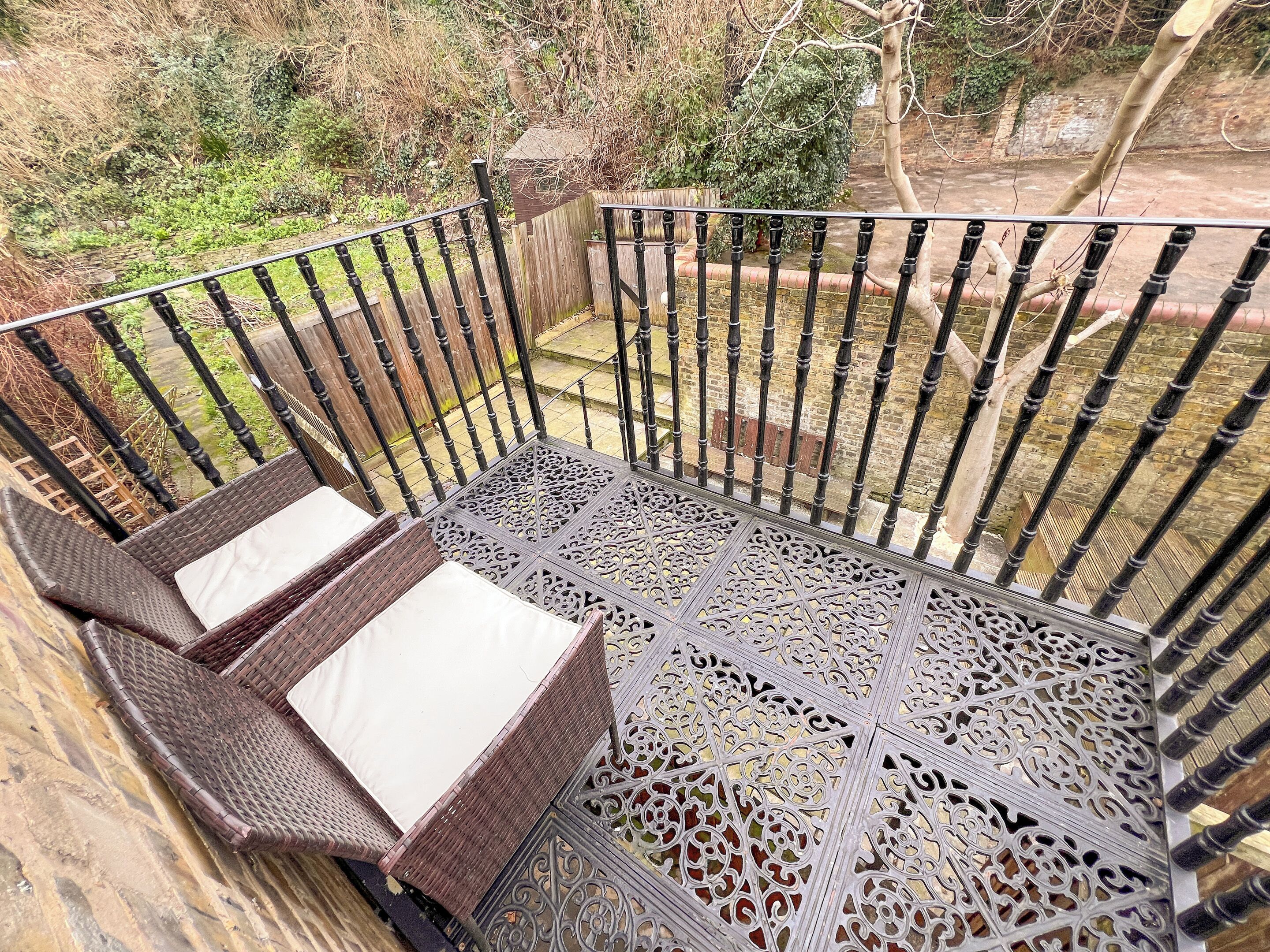 A wicker sofa with cushions on a balcony with ornate metal railings and a view of a garden.