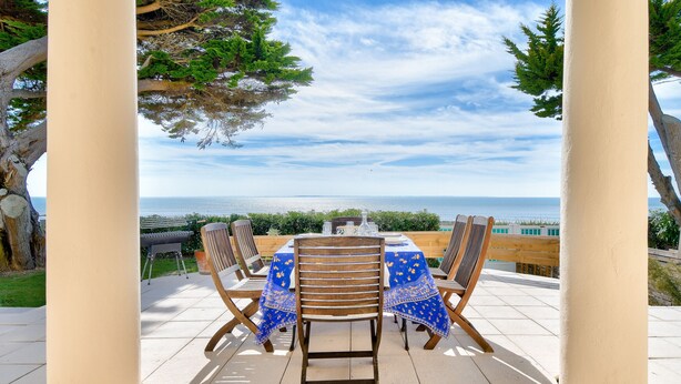 Un patio al aire libre con una mesa dispuesta para cuatro personas, rodeado de sillas de madera, con vistas panorámicas al océano.