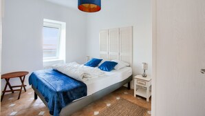 A bedroom with a bed, a nightstand, a lamp, a window, and a hanging light fixture. - Apartment with balcony overlooking the sea, for 4 (Dinard)