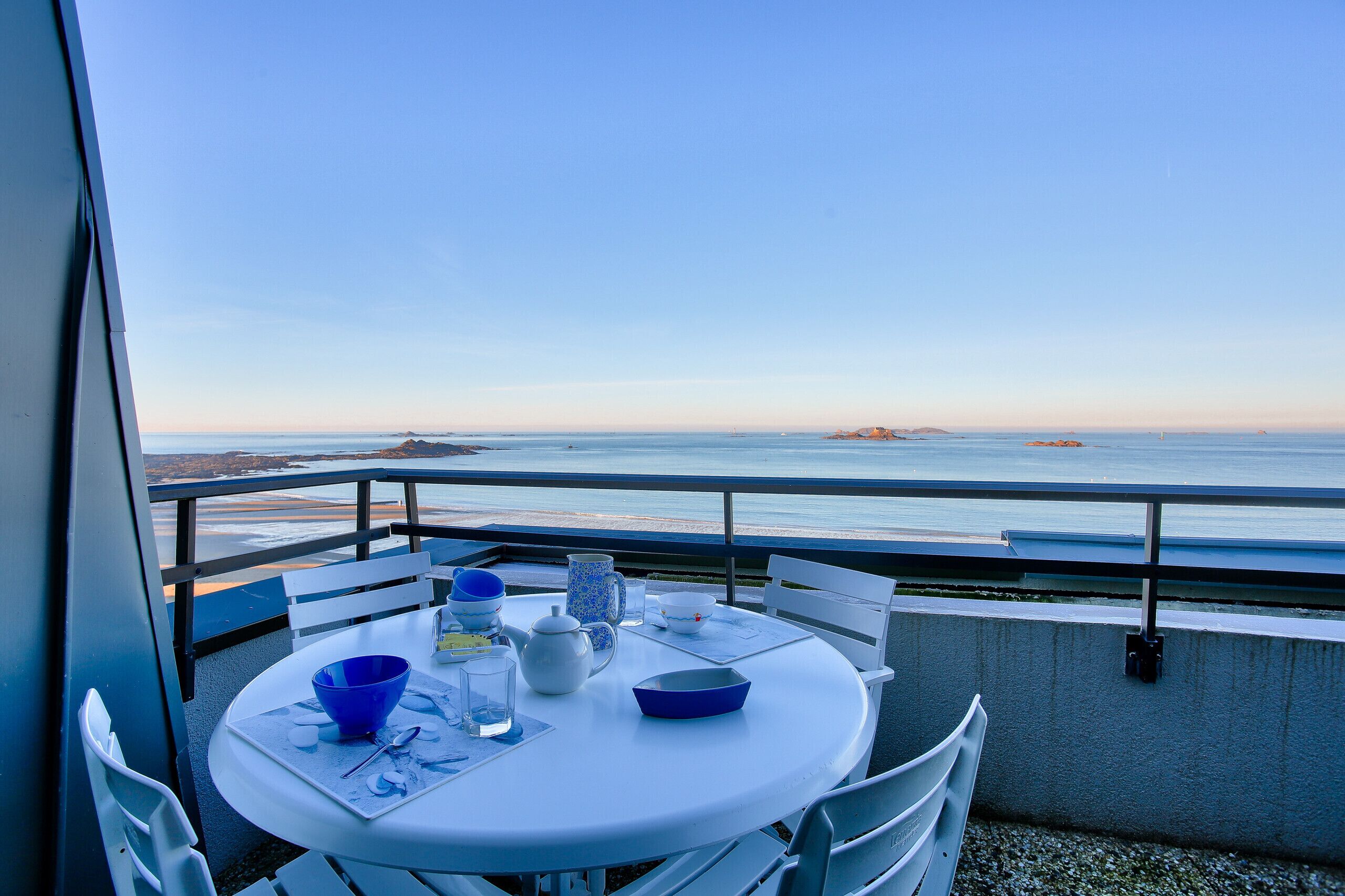 A balcony with a table set for dining, overlooking a calm sea and distant land.