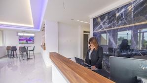 Un área de recepción moderna con un mostrador de madera, una pared de mármol grande y una zona de estar con sillas y una pantalla de televisión.