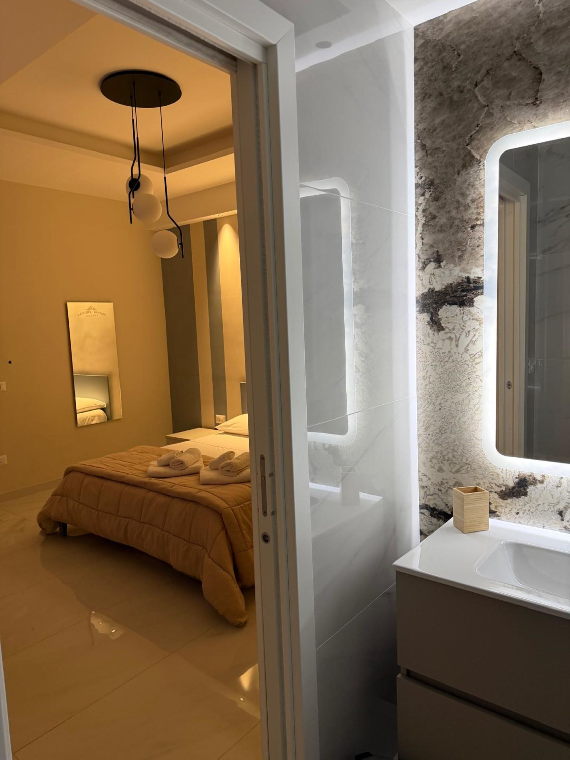 Un dormitorio con cama, mesita de noche, espejo y baño con lavamanos y pared de mármol.