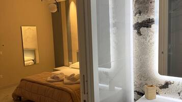 Un dormitorio con cama, mesita de noche, espejo y baño con lavamanos y pared de mármol.