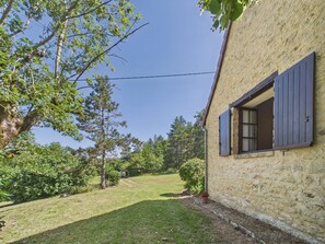 Una casa con fachada de piedra, una ventana con contraventanas y un jardín con árboles y arbustos.