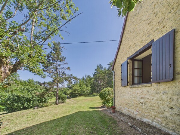 A house with a stone exterior, a window with shutters, and a lawn with trees and shrubs.