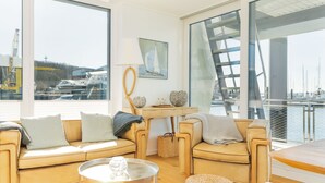 A modern living room with a large window overlooking a marina, a sofa, a coffee table, and a small table with decorative items. - Houseboat mooring FH-LA 02 (Laboe)