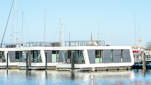 A modern floating structure with multiple decks, docked at a marina with several sailboats in the background. - Houseboat mooring FH-LA 02 (Laboe)